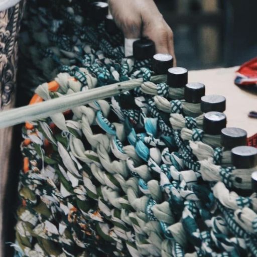 Close-up of a woman weaving a chunky knit blanket on a peg loom using recycled sari fabric, representing therapeutic and dignified work by a survivor artisan.