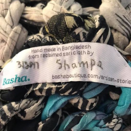 Close-up of a handmade label on a chunky knit blanket, reading "Handmade in Bangladesh from reclaimed sari cloth by Shampa," highlighting the artisan’s name and story.